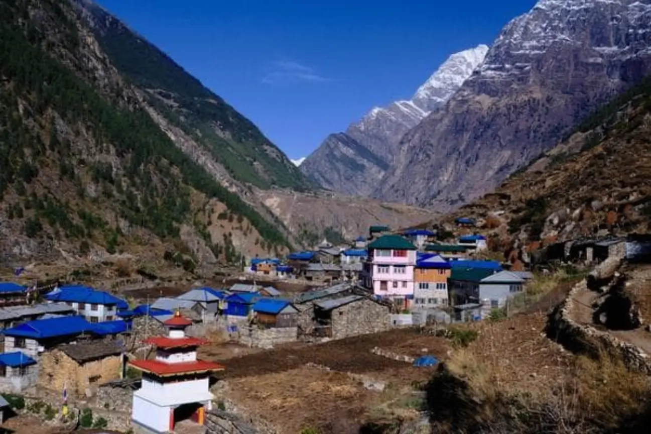 A traditional mountain village with colorful roofs and a white stupa set against steep green hills and snowy peaks for Popular Annapurna Circuit Trek Packages.