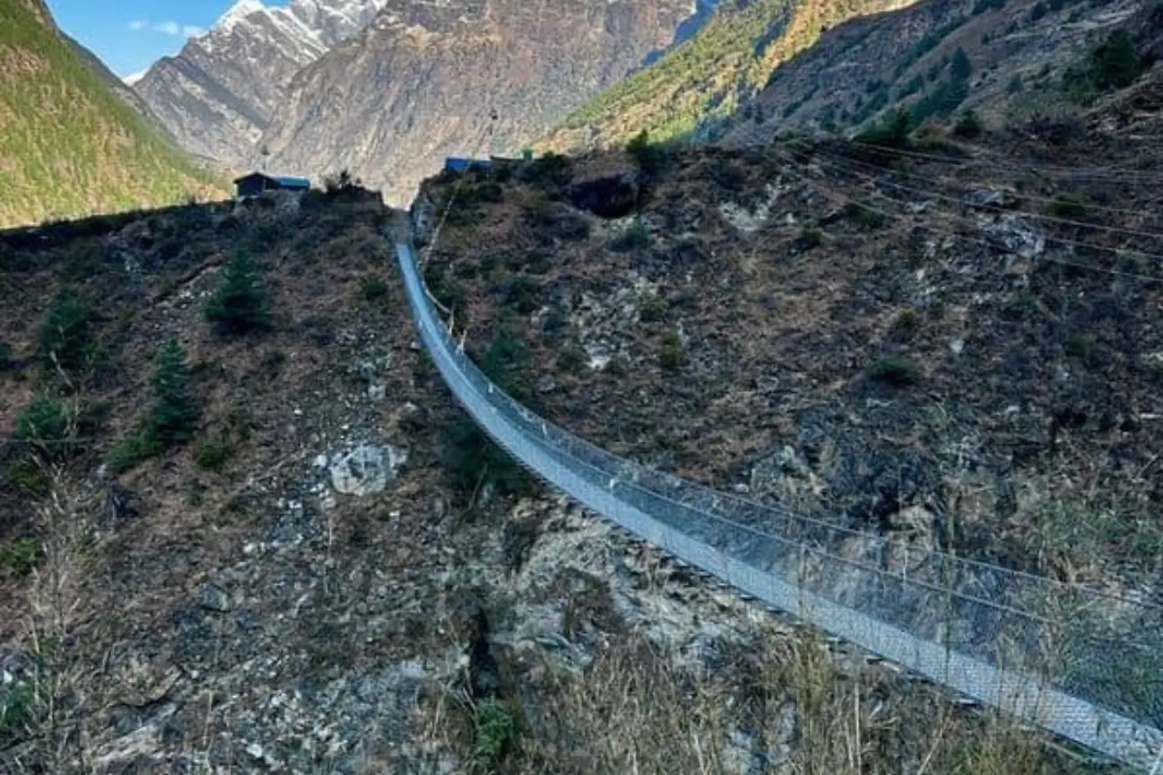 A long steel suspension bridge spanning across a deep canyon with rocky cliffs and distant snowy mountains, featured in Popular Annapurna Circuit Trek Packages.