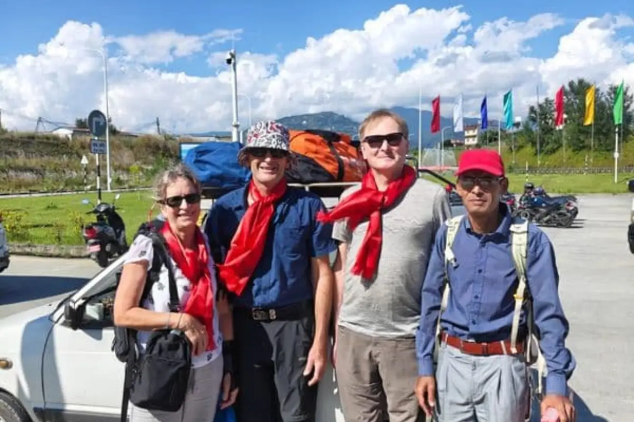 A group of trekkers posing victoriously by the Kang-La Pass sign at 5320 meters with prayer flags and snowy peaks in the background.