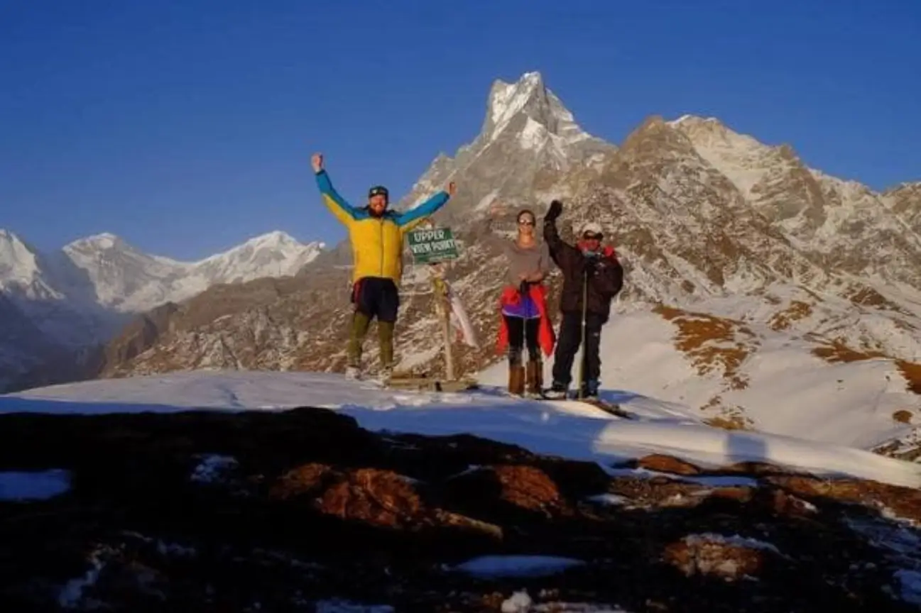 Trekkers celebrating at an upper viewpoint with majestic, snow-covered Himalayan mountain peaks rising sharply in the background.